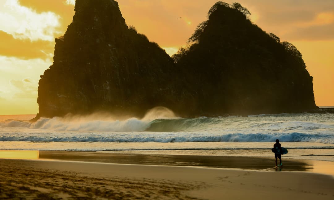 Surfista observando onda tubular ao entardecer diante do Morro Dois Irmãos em Fernando de Noronha